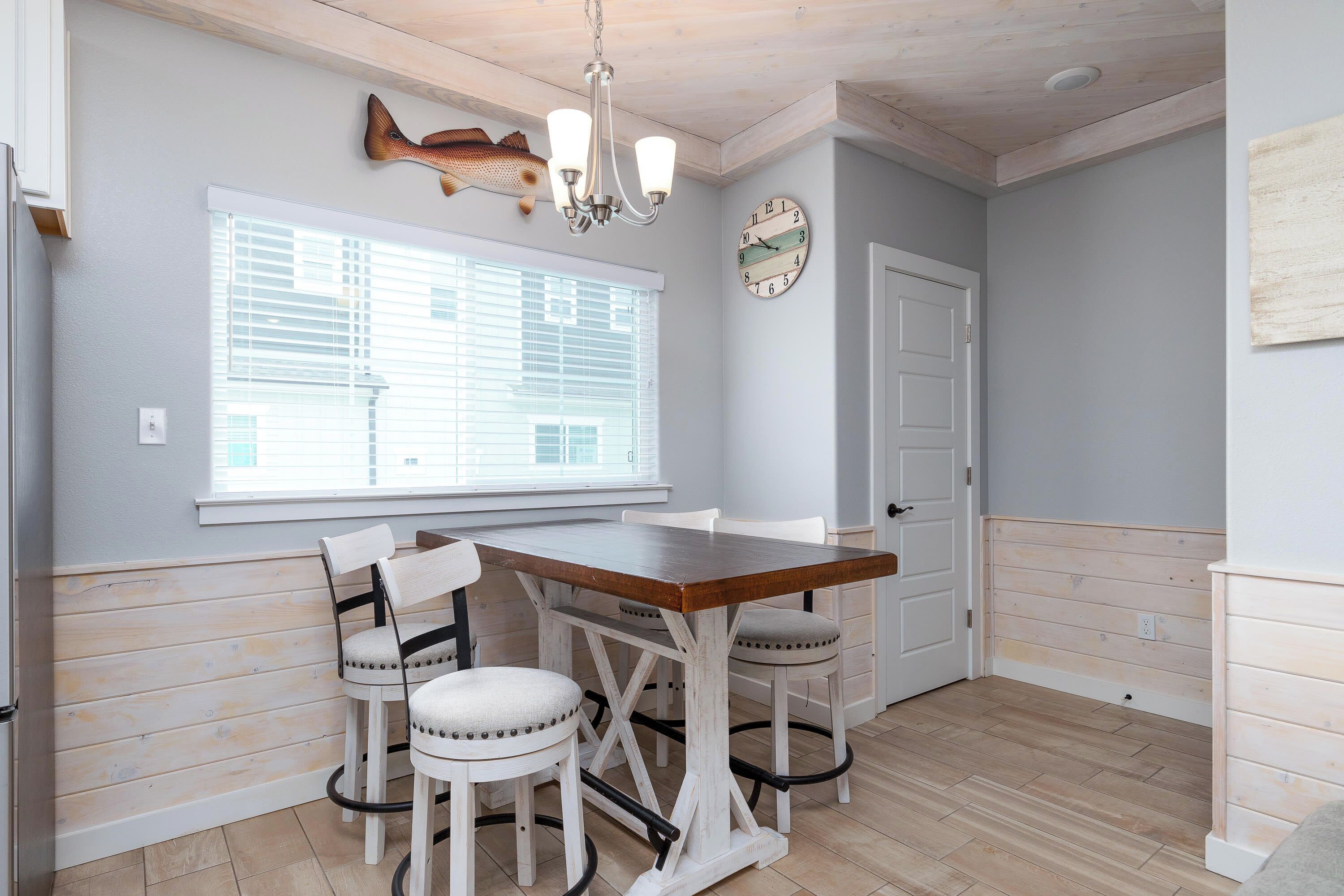 A dining area with a wooden table and chairs, a chandelier, a fish decoration, a clock, and a window with blinds.
