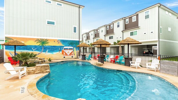 A swimming pool with lounge chairs and umbrellas, surrounded by buildings with a mural of a beach scene. - Modern Comfort, 3 Bedroom, Pets (Port Aransas)