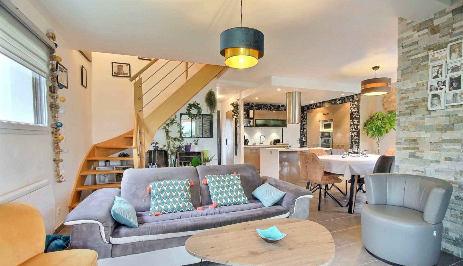 Una sala de estar moderna con sofá, mesa de centro y sillones. Se ve una escalera que sube a una cocina y comedor.