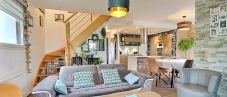 A modern living room with a sofa, coffee table, and armchairs. There is a staircase leading to a kitchen and dining area.