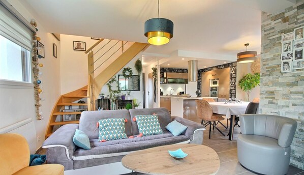 A modern living room with a sofa, coffee table, and armchairs. There is a staircase leading to a kitchen and dining area.