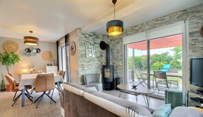A modern living room with a fireplace, dining area, and large windows offering a view of the outdoors.