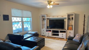 A living room with a large flat-screen TV, leather sofas, a ceiling fan, and a window with blinds.