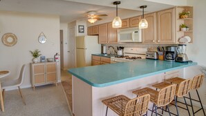 A kitchen with a central island, bar stools, wooden cabinets, a microwave, and various appliances. - Charming Brigantine Condo Rental: Steps to Beach! (Brigantine)