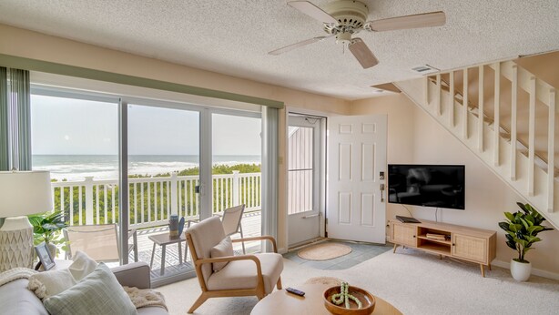 A spacious living room with a large sliding glass door leading to a balcony with a view of the ocean. There is a comfortable sofa, a wooden coffee table, and a television on a wooden stand. - Charming Brigantine Condo Rental: Steps to Beach! (Brigantine)