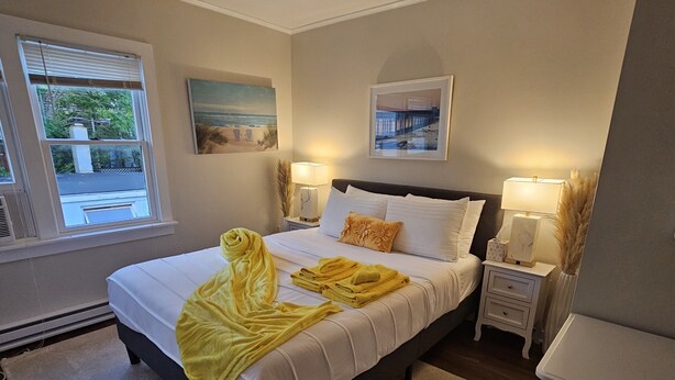 A neatly made bed with white linens and a yellow towel, two bedside lamps, a framed picture of a beach scene, and a window with blinds. - Newly renovated apartment in the heard of Provincetown (Provincetown)