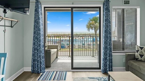 Une pièce dotée d’une grande porte-fenêtre coulissante donnant sur un balcon avec vue sur une piscine et des palmiers.