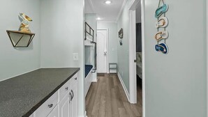 Un couloir étroit avec un coin cuisine sur la gauche, comprenant des meubles blancs et un plan de travail noir. On aperçoit un lit superposé au fond, et une décoration murale représentant un oiseau et une étagère géométrique est accrochée au mur de gauche.