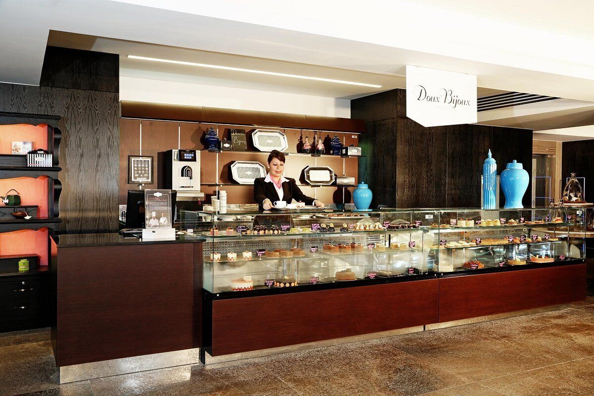 A bakery counter with a variety of pastries and desserts on display, a sign reading "Doux Bijoux," and a person behind the counter.