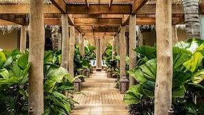 Un sentier en bois, bordé de grandes plantes tropicales et d’lanternes, mène à un espace aménagé avec des sièges, situé sous une pergola en bois.