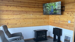 Un accogliente interno di una baita con soffitto in legno, una televisione montata a parete, un mobile intrattenimento nero, un divano grigio e uno specchio con cornice bianca.