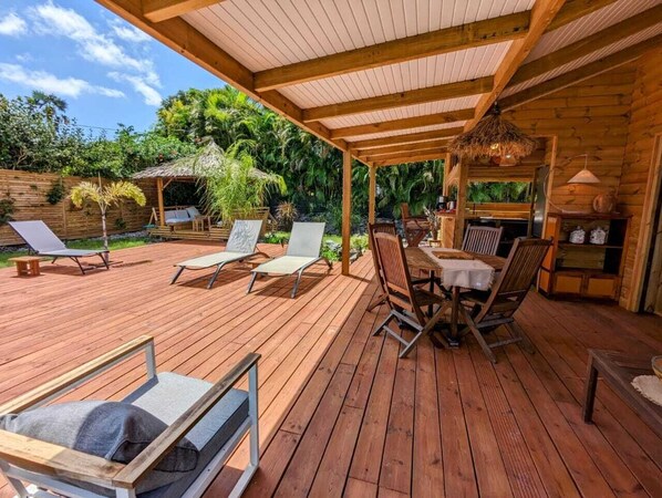 A wooden deck with lounge chairs and a table, surrounded by lush greenery. - Kabane D'o, Gîte Tropical de Charme (Saint-François)