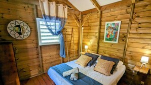 A wooden cabin bedroom with a bed, curtains, a nightstand, and a wall decoration. - Kabane D'o, Gîte Tropical de Charme (Saint-François)