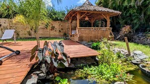A wooden deck with a small pond, surrounded by tropical plants and a gazebo. - Kabane D'o, Gîte Tropical de Charme (Saint-François)