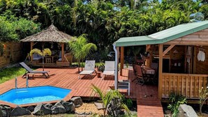 A wooden deck with a swimming pool, lounge chairs, and a covered seating area surrounded by lush greenery. - Kabane D'o, Gîte Tropical de Charme (Saint-François)