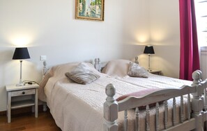 Une chambre à coucher avec un lit double, deux tables de chevet avec des lampes, une commode, un tableau au mur et une fenêtre avec des rideaux.
