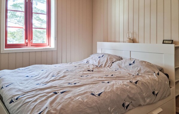 Un lit avec une tête de lit blanche et une couvre-lit à motifs, une fenêtre avec des cadres rouges et un mur avec des lambris verticaux.