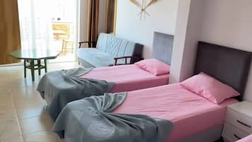 A hotel room with three beds, a small table, a chair, and a view of the outside.