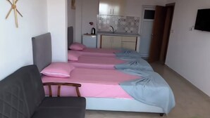 A room with a sofa, a bed with pink and blue bedding, a small table, and a kitchenette in the background. - Avşa apart otel (Marmara)
