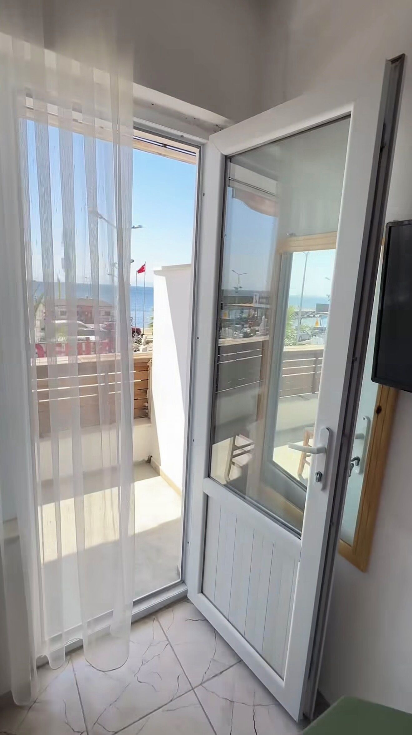 A room with a sliding glass door leading to a balcony with a view of the sea and buildings.