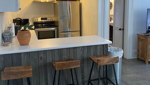 Une cuisine compacte dotée d’un îlot central, d’appareils électroménagers en acier inoxydable et d’un coin bar avec des tabourets en bois.