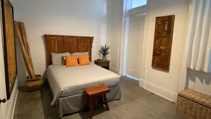 Une chambre à coucher avec un lit, une tête de lit en bois, un petit tabouret en bois, une table de chevet, une plante en pot et un ventilateur de plafond.