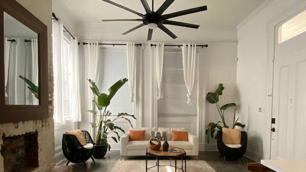 Un salon moderne avec un ventilateur de plafond, des rideaux blancs, un canapé blanc, deux fauteuils noirs, une table basse et une cheminée.