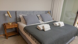 Una cama bien hecha con una colcha gris y dos almohadas, una mesita de noche de madera con una lámpara, y una partición de vidrio al fondo.