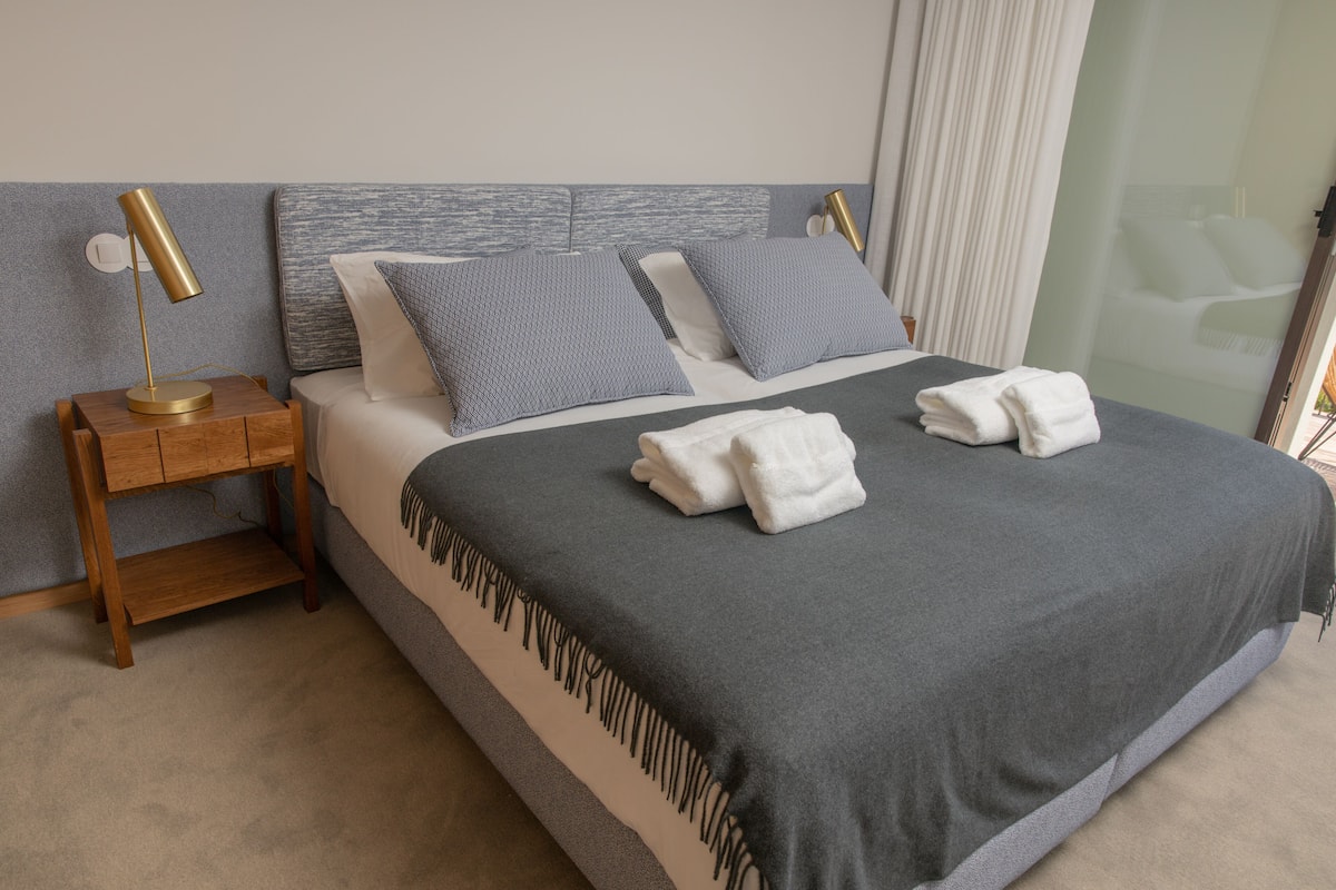 Una cama bien hecha con una colcha gris y dos almohadas, una mesita de noche de madera con una lámpara, y una partición de vidrio al fondo.
