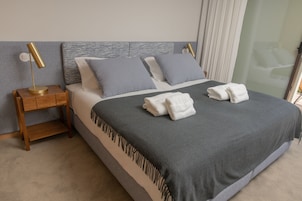 Una cama bien hecha con una colcha gris y dos almohadas, una mesita de noche de madera con una lámpara, y una partición de vidrio al fondo.