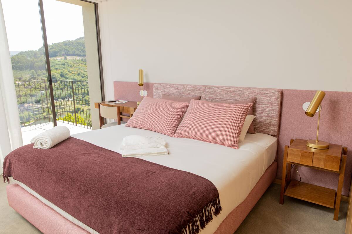 Una cama bien hecha con ropa de cama rosa y blanca, una mesita de noche de madera con una lámpara, y un balcón con vistas a zonas verdes.