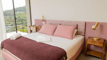 A neatly made bed with pink and white bedding, a wooden nightstand with a lamp, and a balcony with a view of greenery.