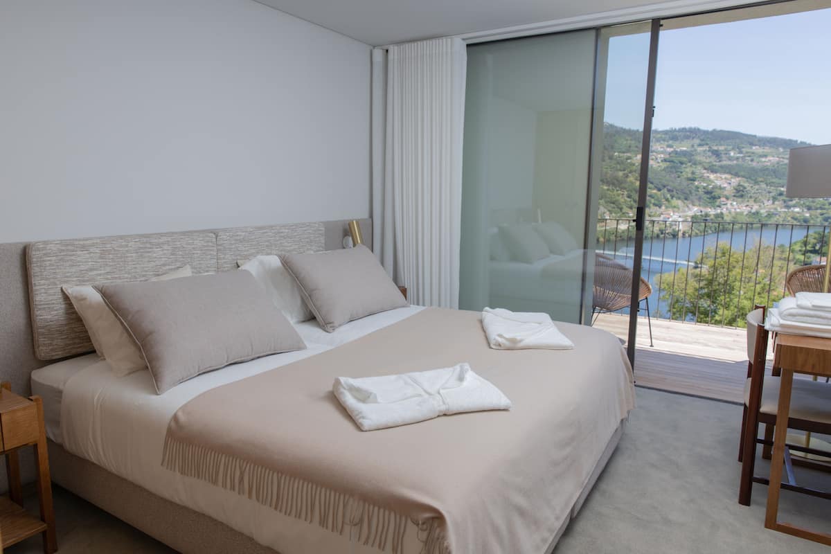 Una habitación de hotel moderna con una cama grande, un balcón con vistas a un lago y colinas, y un comedor con mesa y sillas.