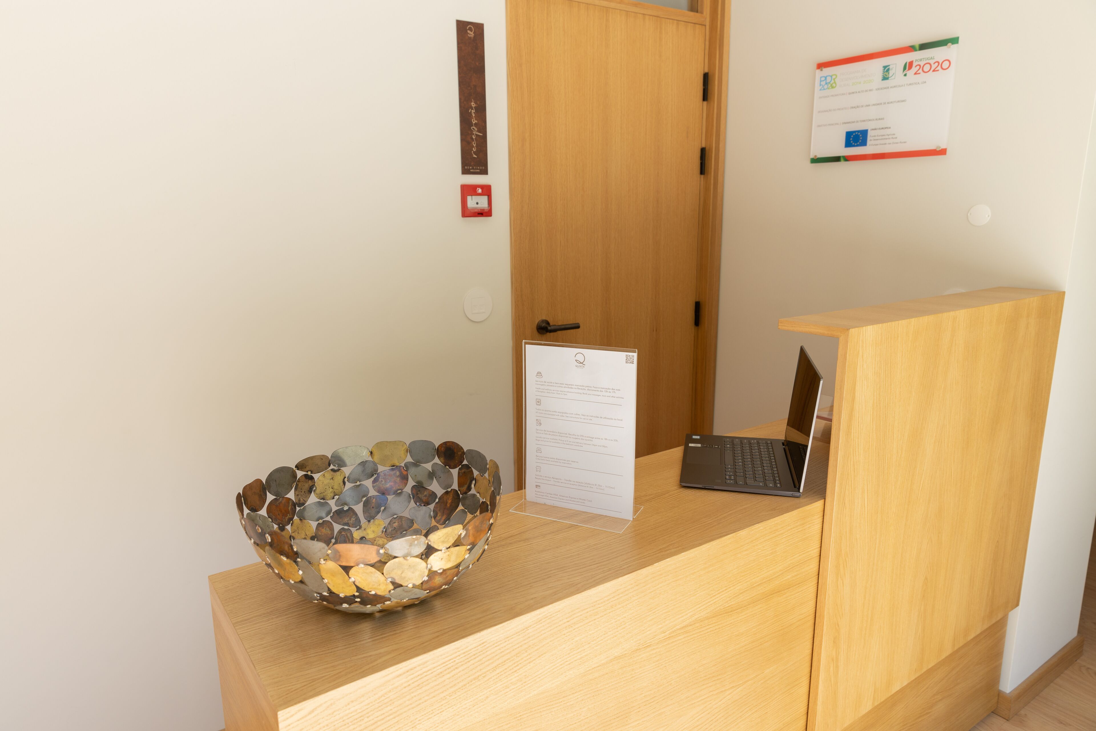 Een receptiebalie met een laptop, een decoratieve glazen schaal, een bord en een houten deur.