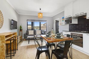 A modern kitchen with white cabinets, a dining table with chairs, a sofa, and a TV mounted on the wall. - Cape on Porterfield 5 (Cape Town)