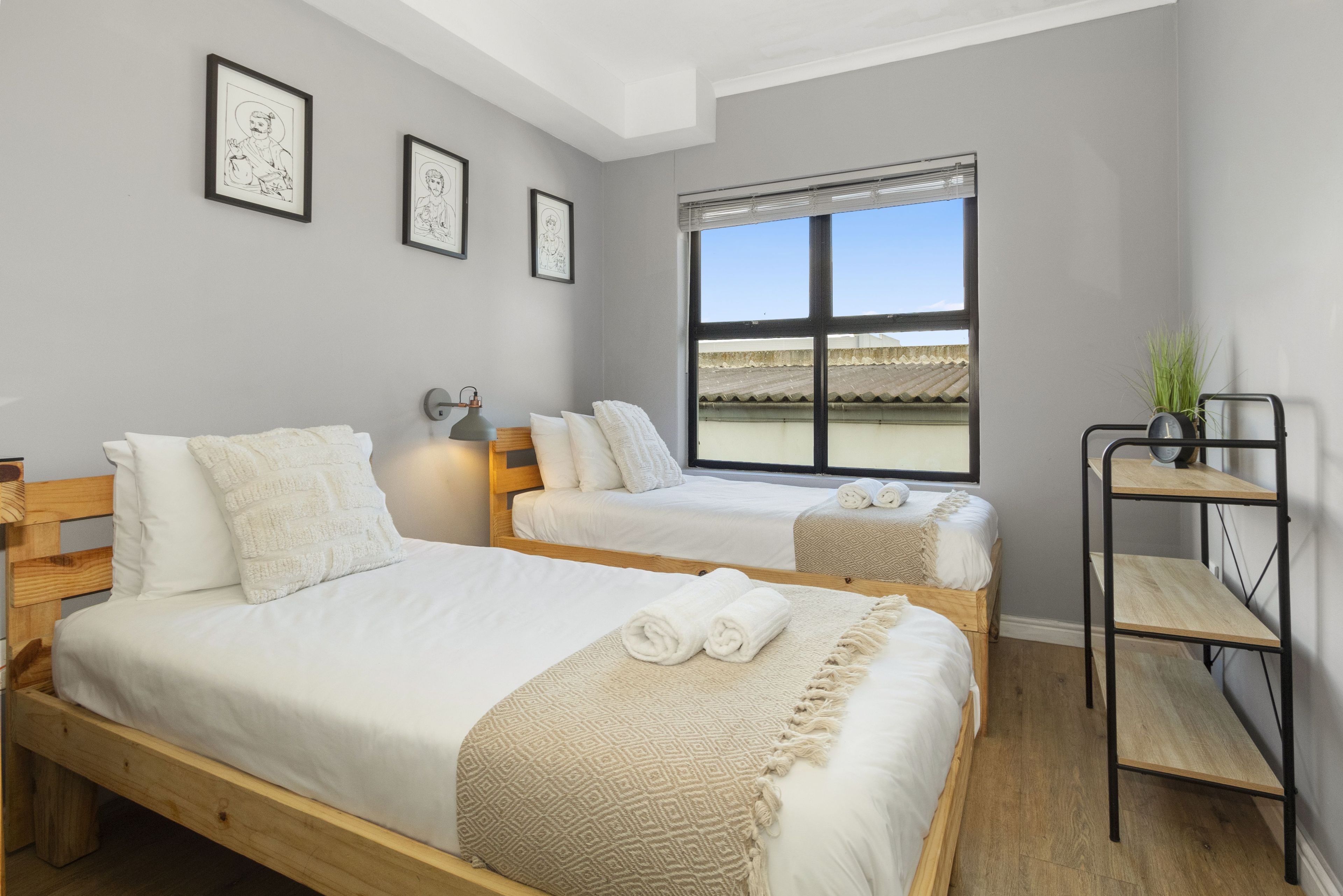 A hotel room with two beds, a window with blinds, a small table with a plant, and framed pictures on the wall.
