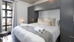 A modern hotel room with a large bed, grey wardrobe, and a view of the city through a large window. - Manhattan on Coral 7 (Cape Town)