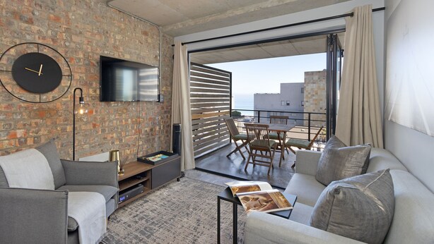 A modern living room with a brick wall, a flat-screen TV, a sofa, a coffee table, and a dining area with a table and chairs. - Manhattan on Coral 7 (Cape Town)