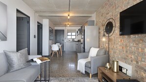 A modern living room with a grey sofa, a coffee table, and a TV mounted on a brick wall. There is a kitchen in the background with a dining area. - Manhattan on Coral 7 (Cape Town)