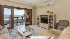 A living room with a glass table, beige sofas, a wooden entertainment unit, and a view of the ocean through a sliding glass door. - Summer Place 9 (Cape Town)