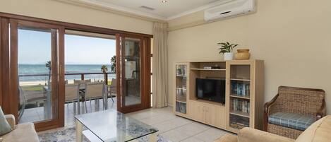 Una sala de estar con una mesa de vidrio, sofás color beige, un mueble de entretenimiento de madera y vista al océano a través de una puerta corrediza de vidrio.