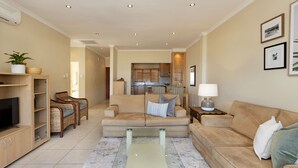 A living room with a beige sofa, a glass coffee table, wicker chairs, a TV, and a kitchen in the background. - Summer Place 9 (Cape Town)