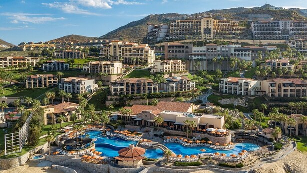 Un resort con varias edificaciones, una piscina central y un paisaje montañoso de fondo.