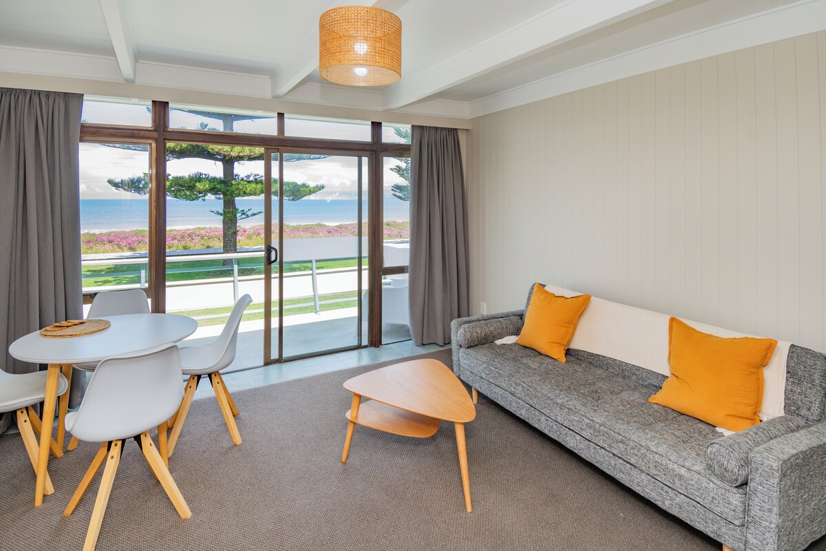 Ein modernes Wohnzimmer mit einem grauen Sofa, zwei orangefarbenen Kissen, einem runden weißen Tisch mit Stühlen und Blick auf das Meer durch große Fenster.