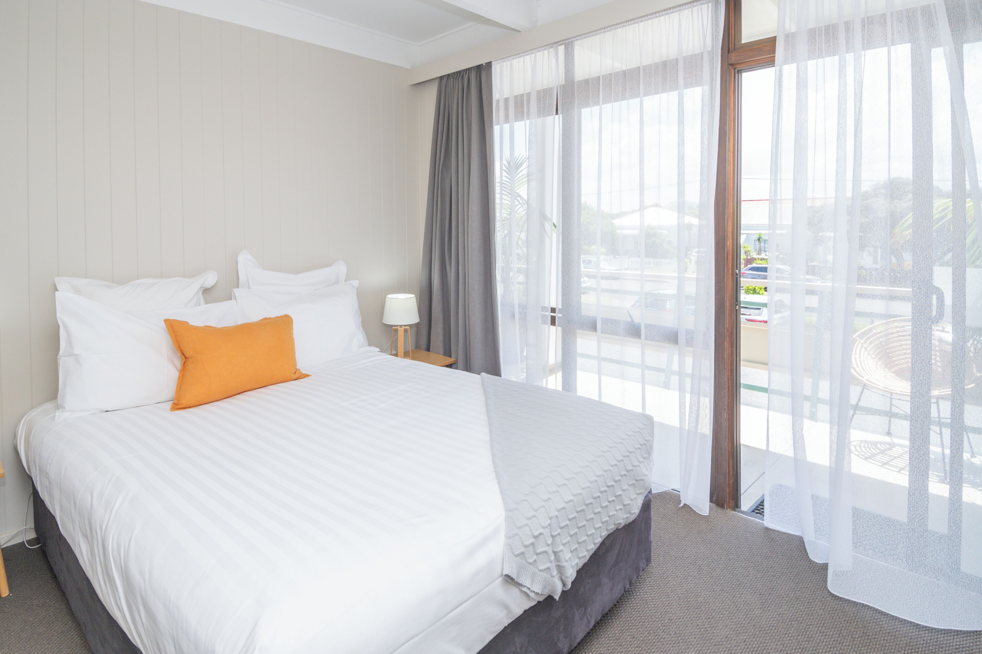 A hotel room with a large bed, a nightstand, a lamp, and a view of the outside.