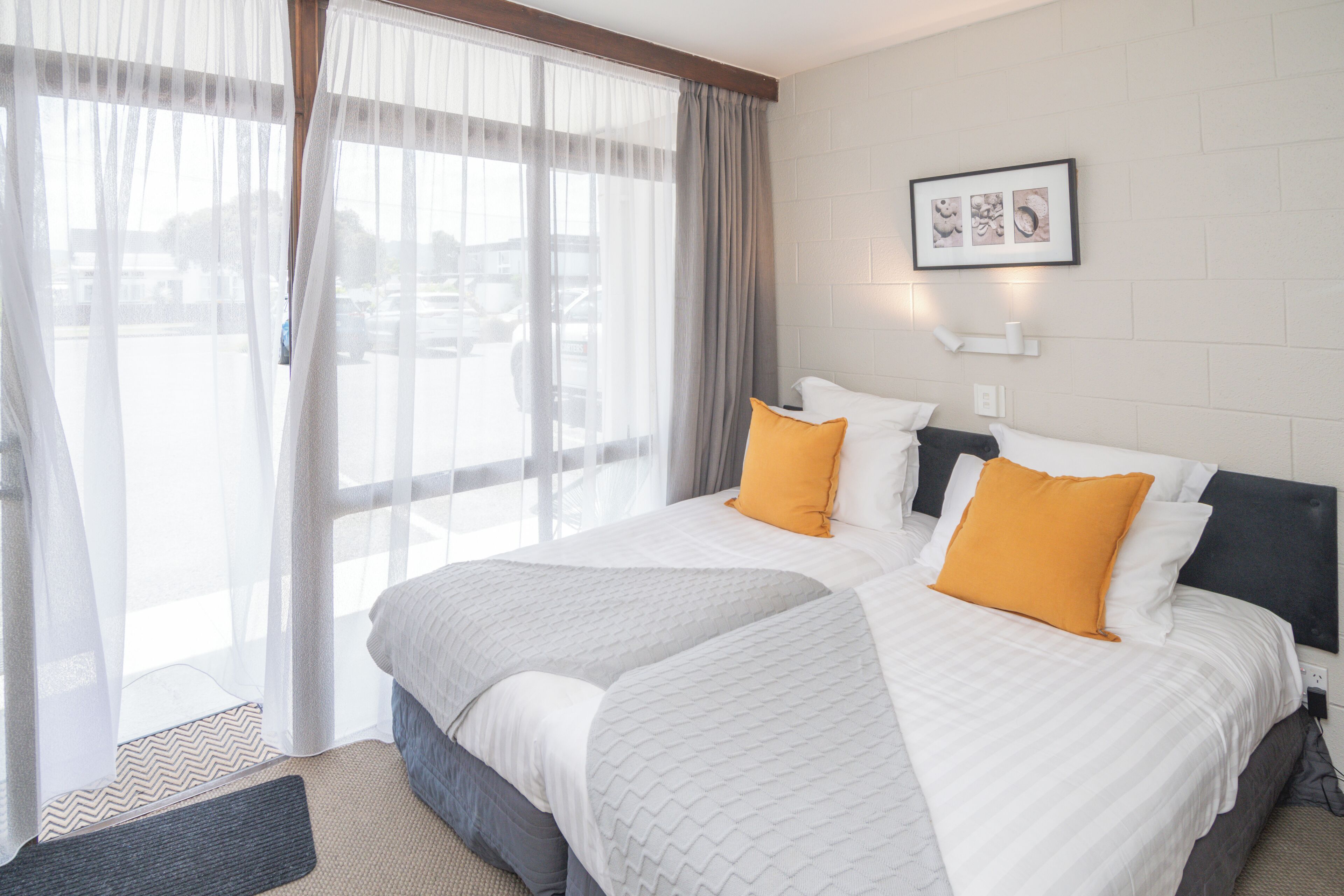 A hotel room with two beds, a large window with sheer curtains, a framed picture on the wall, and a rug on the floor.