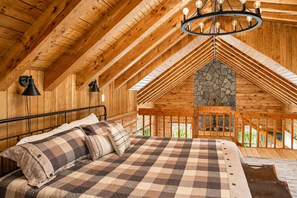 A cozy bedroom with a checkered bedspread, a stone fireplace, and a wooden ceiling.