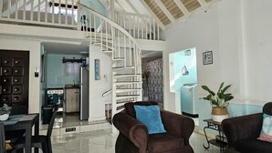 Een ruime woonkamer met een hoge plafond, een witte trap, een eethoek met een tafel en stoelen, en een keuken op de achtergrond.
