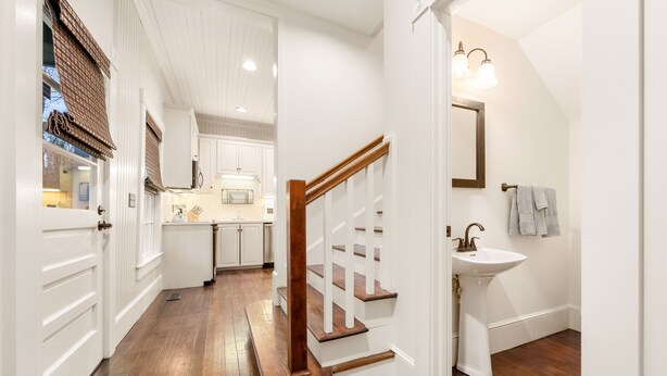 Un pasillo con piso de madera, una escalera blanca y una zona de cocina al fondo.