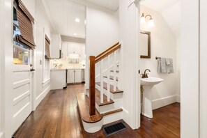 Un pasillo con piso de madera, una escalera blanca y una zona de cocina al fondo.